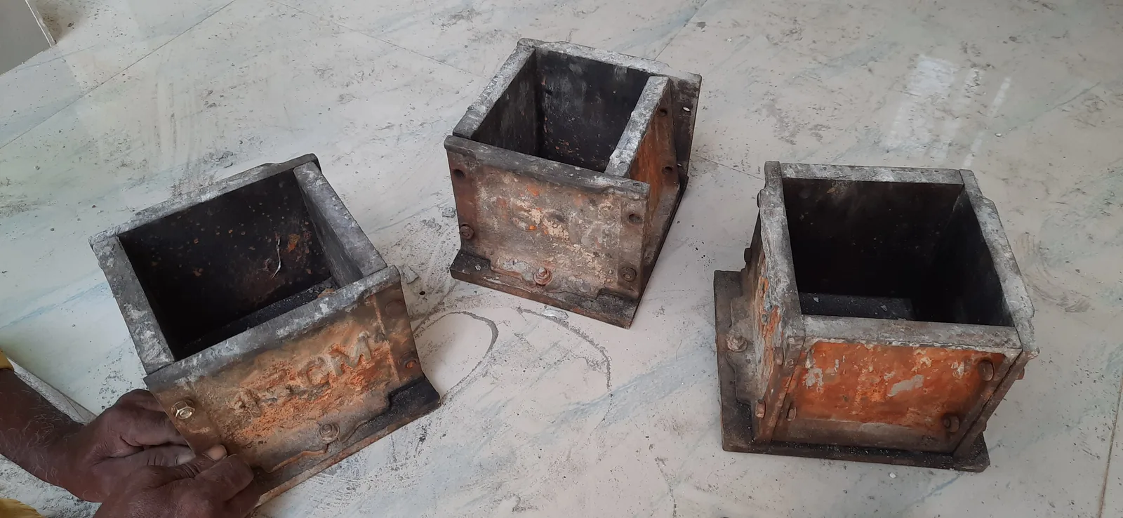 Three empty steel moulds for concrete test cubes standing on a dusty floor