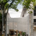 Street view of a translucent perforated metal facade set back behind a paved forecourt under a large tree