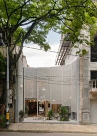 Street view of a translucent perforated metal facade set back behind a paved forecourt under a large tree