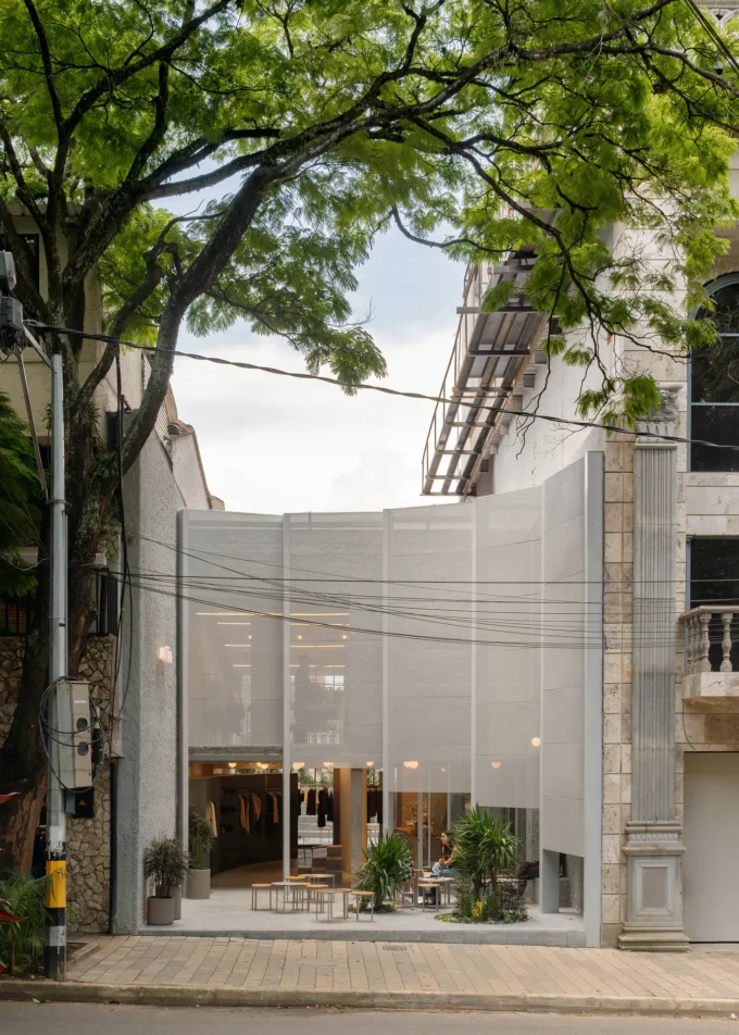 Street view of a translucent perforated metal facade set back behind a paved forecourt under a large tree