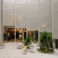 Forecourt with round café tables, palm planters and the glowing shop behind the mesh screen