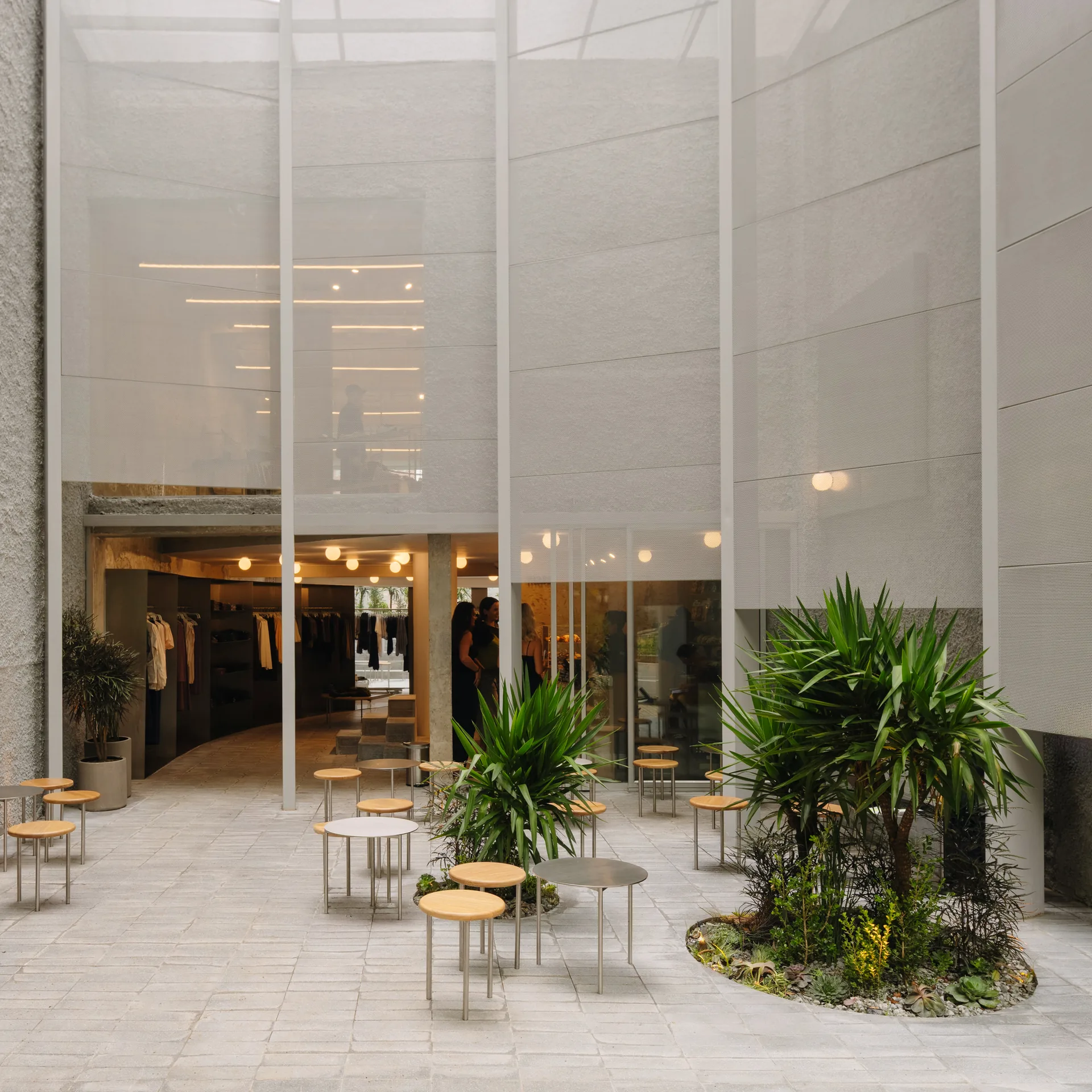 Forecourt with round café tables, palm planters and the glowing shop behind the mesh screen