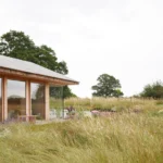 Low barn with a timber shingled pitched roof, tall glazing and a pale chimney seen across a wildflower meadow, The Makers Barn by HUTCH design
