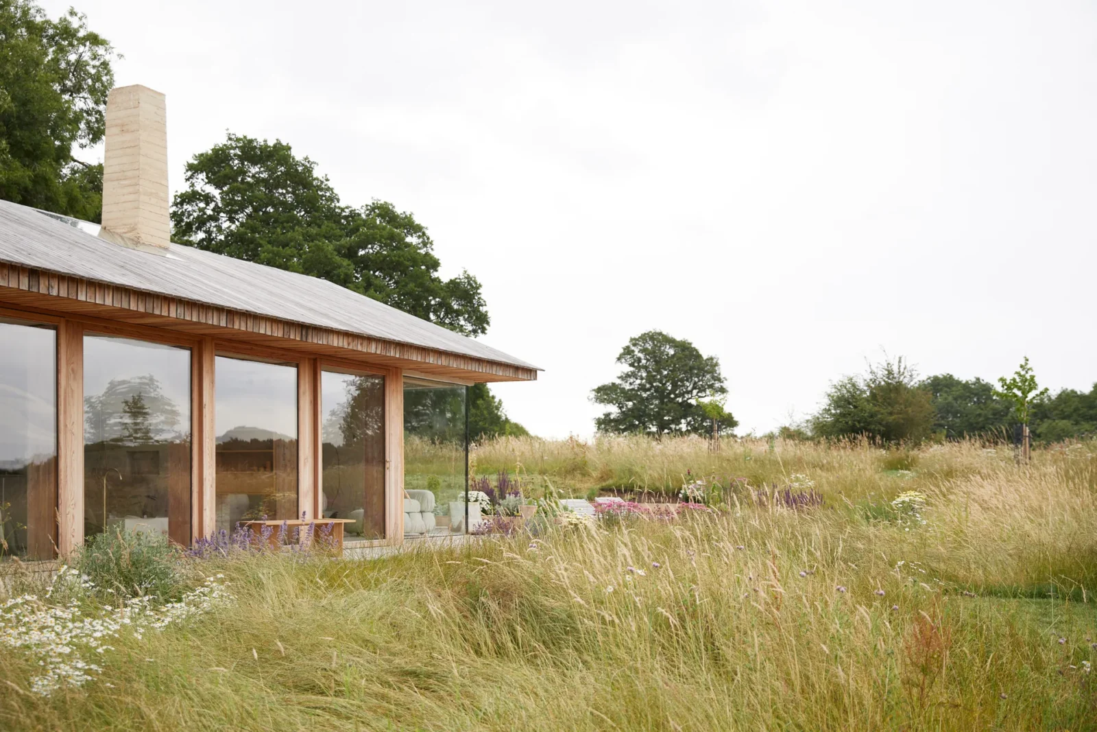 Low barn with a timber shingled pitched roof, tall glazing and a pale chimney seen across a wildflower meadow, The Makers Barn by HUTCH design