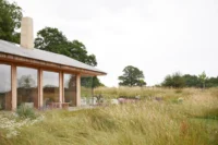 Low barn with a timber shingled pitched roof, tall glazing and a pale chimney seen across a wildflower meadow, The Makers Barn by HUTCH design