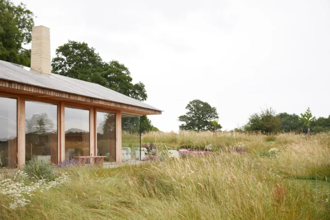 Low barn with a timber shingled pitched roof, tall glazing and a pale chimney seen across a wildflower meadow, The Makers Barn by HUTCH design