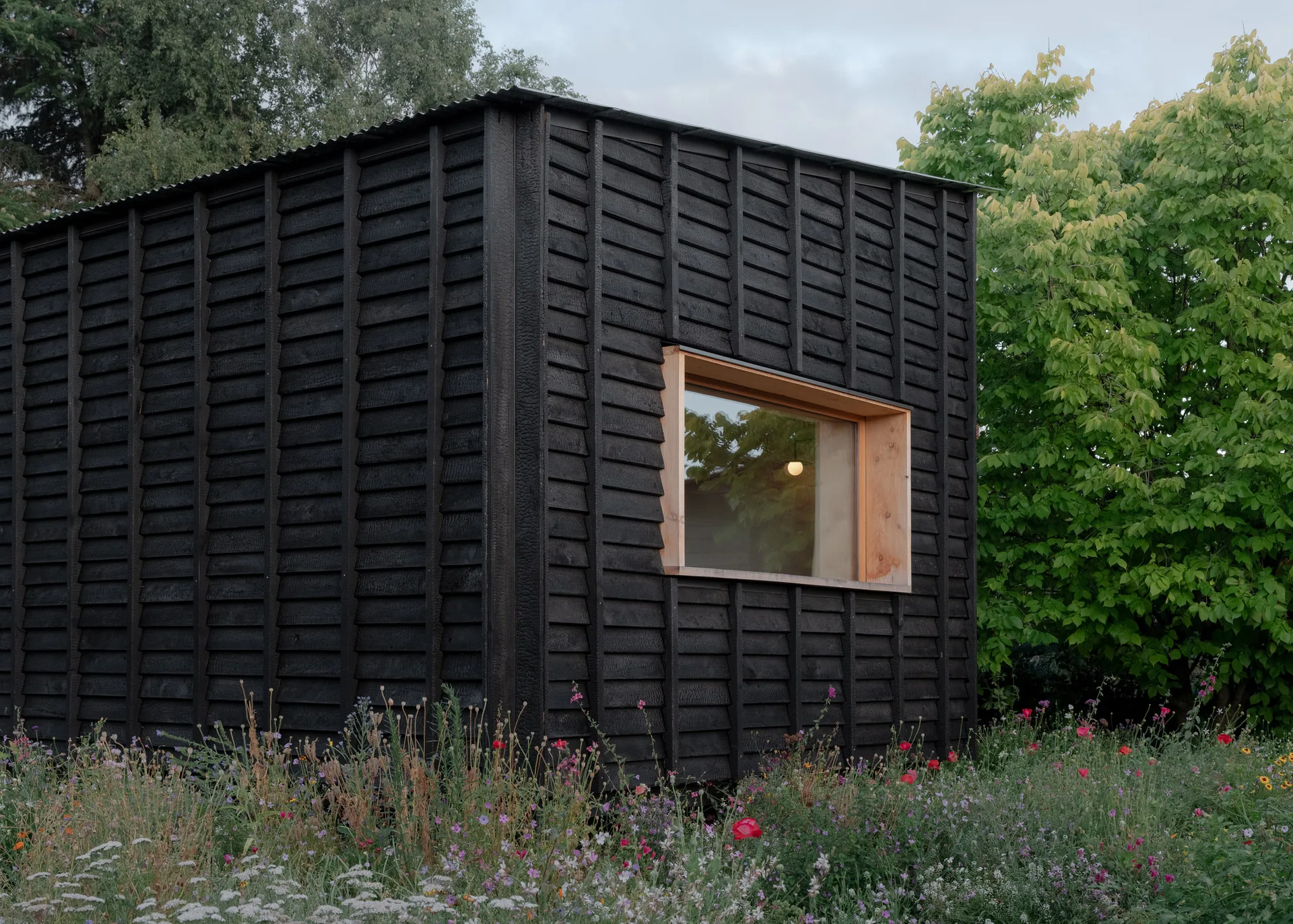Small black charred timber cabin with a deep timber framed window set in a wildflower garden, The Yakisugi Cabin