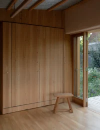 Wall of pale timber cupboards with a stool beside a glazed door in the main room, The Yakisugi Cabin