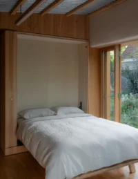 Fold down bed revealed inside the timber wardrobe wall beside a glazed door, The Yakisugi Cabin