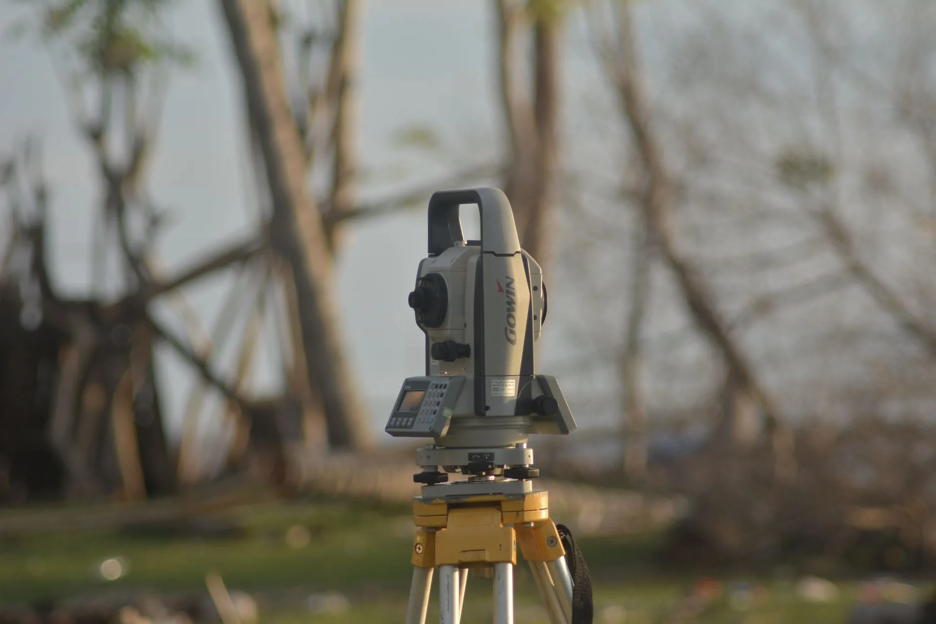 Surveying total station mounted on a yellow tripod outdoors in front of blurred trees