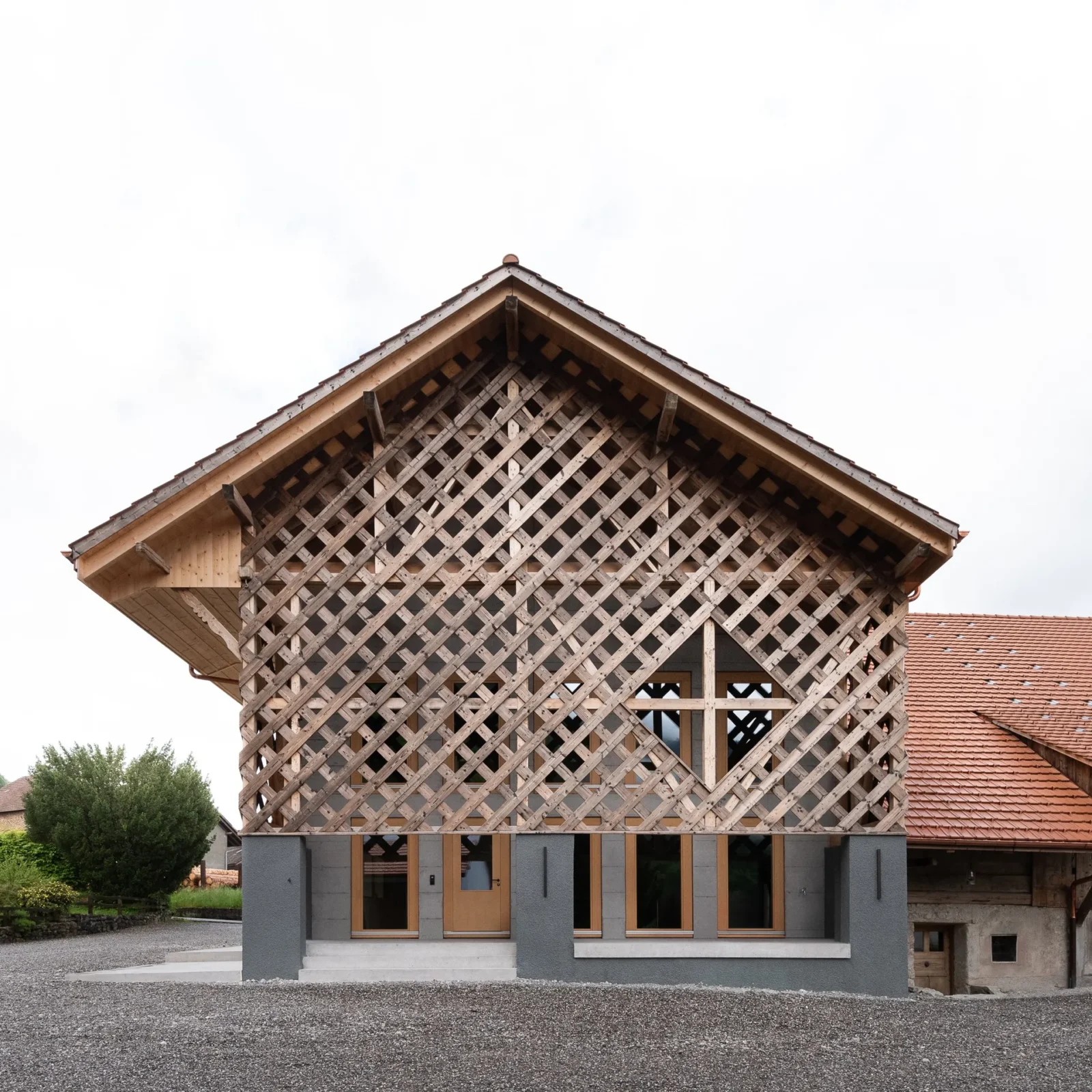 Gable end of the farmhouse rebuilt as a diagonal timber lattice above grey concrete piers and glazed doors