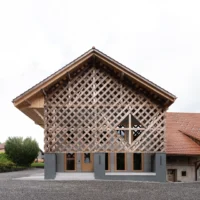 Gable end of the farmhouse rebuilt as a diagonal timber lattice above grey concrete piers and glazed doors