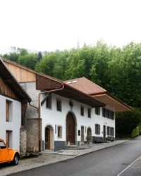 Farmhouse seen along the village road with a stone arched door, an orange car and wooded hills behind