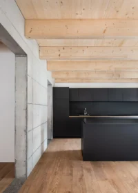 Black kitchen with an island beside a concrete wall under a timber beamed ceiling