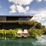 Dark two storey house draped in plants behind an infinity pool and sun loungers, Vista House by Studio Gontijo Arquitetura